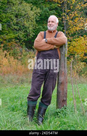 Im Landhausstil mit Latzhose, Pullover und Gummistiefeln lehnt sich ein älterer Mann auf der Wiese an einen Holzpfosten. Stockfoto