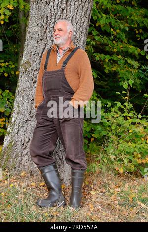 Lächelnder älterer Mann im lässigen Landhausstil mit braunen Latzhose, Pullover und Gummistiefeln lehnt sich an einen Baumstamm am Waldrand. Stockfoto