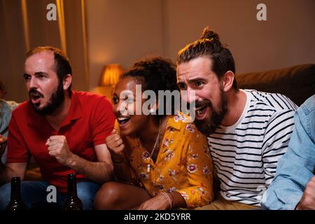 Eine Gruppe von drei jungen Freunden, die zu Hause auf der Couch sitzen und gemeinsam Sport im fernsehen gucken, schreiend fröhlich. Sportkonzept. Stockfoto