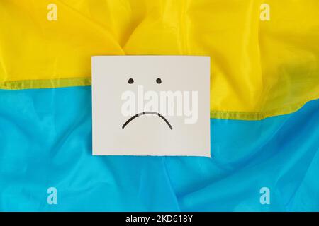 Auf den Flaggen der Ukraine liegt ein trauriges, auf Papier gezeichnetes Emoticon, eine schlechte Laune Stockfoto