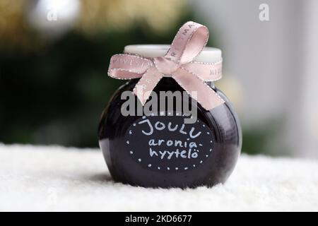 Hausgemachte weihnachtliche Marmelade in einem Glas mit hellroter Schleife. Apfelbeermarmelade im Inneren. Nahaufnahme des Farbbildes mit dem Baum im Hintergrund. Stockfoto