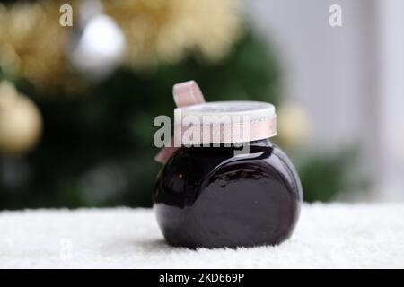 Hausgemachte weihnachtliche Marmelade in einem Glas mit hellroter Schleife. Apfelbeermarmelade im Inneren. Nahaufnahme des Farbbildes mit dem Baum im Hintergrund. Stockfoto