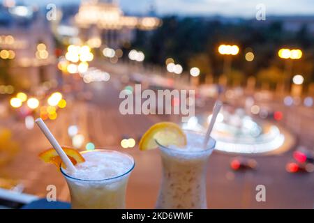 Cocktails mit einem Strohhalm und einem Stück Zitrone auf einem Glas. Verschwommene Stadt mit Laternen im Hintergrund. Ein Drink für zwei, romantische Atmosphäre, Date für Stockfoto