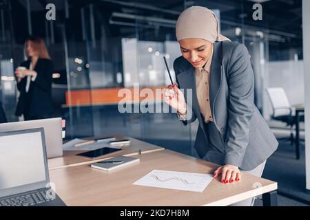 Lächelnde muslimische Geschäftsfrau im Hijab, die mit Dokumenten arbeitet und Seite anschaut Stockfoto