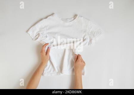 Ein weißes T-Shirt liegt auf einem weißen Hintergrund und weibliche Hände, Damenmode Kleidung Stockfoto