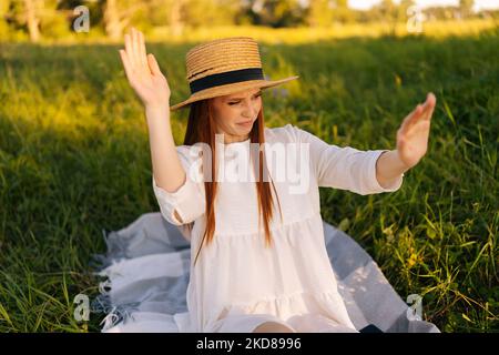 Porträt der unzufriedenen Rotschopf hübsche Frau in Strohhut und weißem Kleid jagen Insekten weg von sich selbst sitzen auf dem Feld mit grünem Gras. Stockfoto