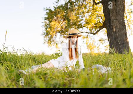 Porträt der eleganten Rotschopf junge Frau in Strohhut und weißem Kleid arbeiten auf Laptop-Computer suchen, um anzuzeigen, sitzen auf dem Feld mit grünem Gras. Stockfoto