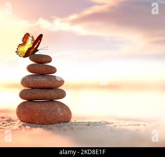 Ausgewogene Kieselsteinpyramide und Schmetterling am Strand. Abstrakt warmer Sonnenuntergang Bokeh mit Sand auf dem Hintergrund. Zen-Steine am Meeresstrand, Balance Conce Stockfoto
