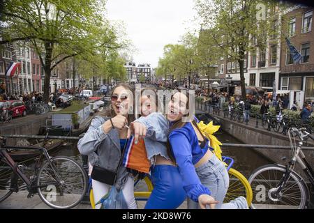 Drei junge holländische Mädchen mit der holländischen Flagge auf ihren Gesichtern posieren fröhlich vor dem Kanal. Die Niederlande feiern den Königstag nach zwei Jahren der Annullierung aufgrund der COVID-19 Coronavirus-Pandemie und der Sperrbeschränkungen und -Maßnahmen. Tausende von Einheimischen, Feiernden und Besuchern haben das Stadtzentrum, die Parks in der Nähe des Rijksmuseums, die Grachten von Amsterdam besucht, um mit verschiedenen Festlichkeiten den Geburtstag von König Willem-Alexander zu feiern, der als Koningsdag bekannt ist, ein niederländischer Nationalfeiertag. Alle Arten von Booten und Schiffen sind in den Kanälen unter den berühmten Brücken und in der Nähe Th vorbei gesehen Stockfoto