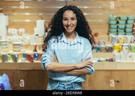 Portrait of business owner, hispanic woman managing gift shop, woman with curly hair looking at camera and smiling with arms crossed. Stockfoto