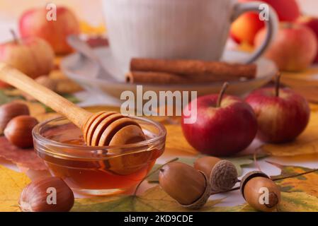 Weißer Tisch mit Herbstschmuck, einer Tasse Tee, Zimtstücken, einer Glasschale mit Honig, kleinen Äpfeln, Nüssen, bunten Baumblättern und Eicheln. Niedrig Stockfoto