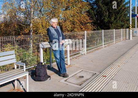 Ein alter grauhaariger gelangweilter Tourist in blauen Jeans, der sich auf einer modernen Holzbank oder einem lehnenden Stand oder einer schlanken Bar (noch kein Name) lehnte und mit einem Rücken wartete Stockfoto