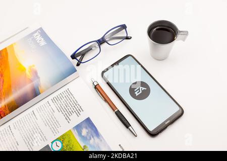 In dieser Fotolillustration ist ein EA-Logo (Electronic Arts Sports) zu sehen, das am 31. Mai 2022 auf einem Smartphone-Bildschirm auf einem Schreibtisch neben einem Café, einem Stift, einer Brille und einem Magazin in Athen, Griechenland, zu sehen ist. (Foto-Illustration von Nikolas Kokovlis/NurPhoto) Stockfoto