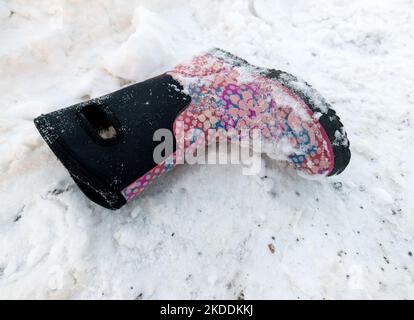 Ein leerer Winterstiefel für Kinder, der auf Schnee liegt. Der Stiefel ist rosa, rot, blau und schwarz. Es liegt Schnee darauf. Stockfoto