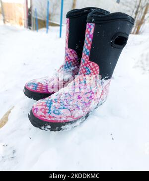 Ein leeres Paar Winterstiefel für Kinder, die auf einer Schneebank ruhen. Die Stiefel sind rosa, rot, blau und schwarz. Es liegt Schnee auf ihnen. Stockfoto