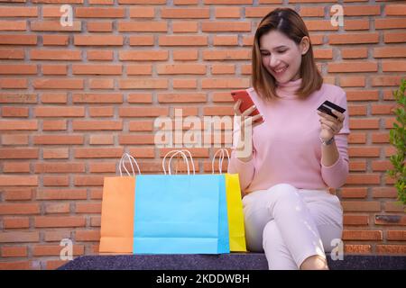 Junge Frau kauft online mit Smartphone und Kreditkarte mit bunten Einkaufstaschen daneben. Stockfoto