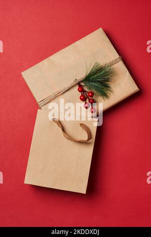 Ein weihnachtsgeschenk, eingewickelt in braunes Papier mit roten Beeren und grünen Blättern, das auf einem roten Hintergrund sitzt Stockfoto