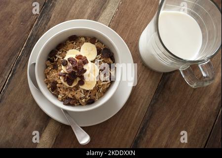 Top down First Person Perspective Blick auf eine kleine weiße Schüssel Müsli mit Joghurt, Rosinen und Bananen auf dem Tisch neben einem Krug mit Milch Stockfoto