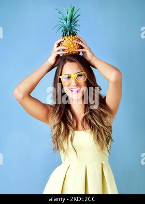 Hallo Sommer, ich bin bereit für dich. Studioaufnahme einer jungen Frau, die auf ihrem Kopf eine Ananas vor blauem Hintergrund balanciert. Stockfoto