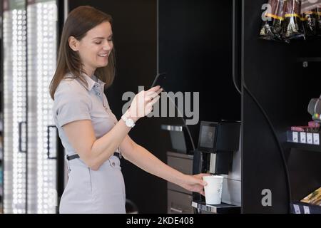 Geschäftsfrau hält Smartphone und bereitet Kaffee mit einem professionellen Verkaufsautomaten zu Stockfoto