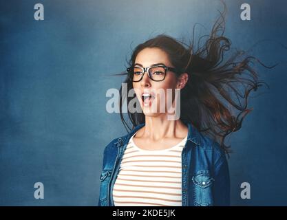 Umgehauen. Studioaufnahme einer jungen Frau mit Luft, die vor blauem Hintergrund in ihr Gesicht geblasen wird. Stockfoto