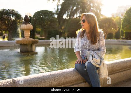 Frau, die am Rand eines Brunnens in einem öffentlichen Park sitzt und die Sonnenstrahlen die Szene von hinten erhellen. Stockfoto