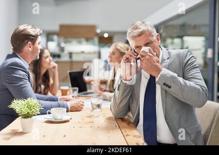 Warten Sie nicht, bis Sie sich schlechter fühlen, bevor Sie den Arzt sehen. Ein Geschäftsmann bläst seine Nase, während er in einem Büro auf einem Mobiltelefon spricht. Stockfoto