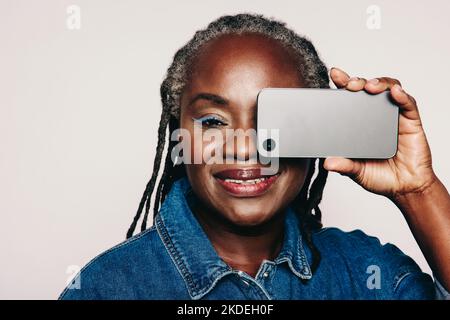 Reife Frau, die auf die Kamera schaut und dabei ein Smartphone vor ihr Auge hält. Modische Frau mit Dreadlocks in Jeansjacke und Make-U Stockfoto