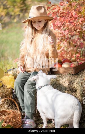 Glückliches Kind Mädchen mit weißem Hund spielt kleinen Landwirt im Herbstgarten Stockfoto