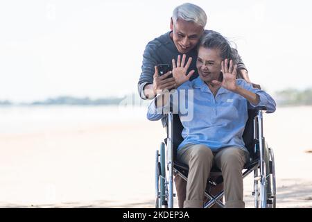 Ältere Frau im Rollstuhl sitzt und Ehemann ist ein Rollstuhl Benutzer Smartphone macht Selfie am Strand Stockfoto