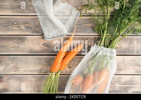 Ein Haufen frischer, reifer Karotten mit langem grünem Schlemm auf einem verblassten Holztisch, Draufsicht. Auspacken von Gemüse aus transparenten Stoffbeuteln. Bio-Lebensmittel oder Stockfoto