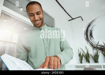 Lächelnder afrikanischer Geschäftsmann, der im modernen Büro steht und Buch hält Stockfoto