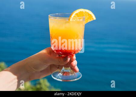 Leicht alkoholisches Getränk mit einer Scheibe Orange in den Händen Frau vor dem Hintergrund des blauen Meeres. Stockfoto
