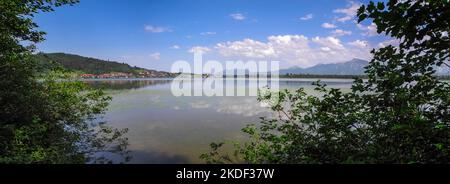 Landschaft Panoramablick auf den Hopfensee am gegenüberliegenden Ufer und das Dorf Hopfen am See vor blauem Himmel mit kleinen weißen Wolken. Stockfoto