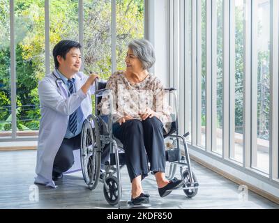 Lächelnd glücklich asiatische ältere weibliche Patientin im Rollstuhl sitzend im Gespräch mit einem freundlichen Mann Arzt im weißen Mantel in der medizinischen Praxis im Krankenhaus. Stockfoto