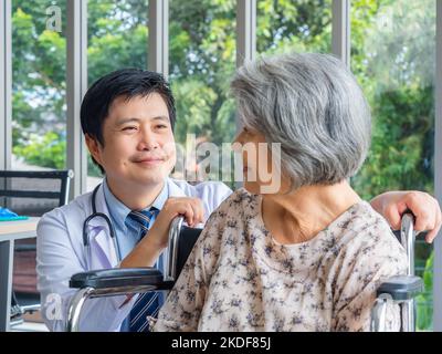 Lächelnd positiver asiatischer Mann Arzt im weißen Mantel spricht mit Sorgfalt zu älteren älteren älteren weiblichen Patienten, die in einem Rollstuhl in der medizinischen Praxis in der Zusammenarbeit sitzen Stockfoto