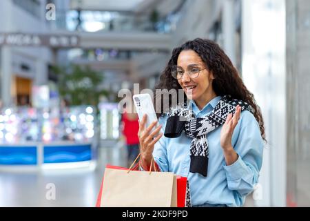 Glückliche weibliche Shopper im Laden, die mit Freunden über Videoanruf sprechen, hispanische Frau, die Smartphone für die Online-Fernkommunikation nutzt, Informationen über Verkäufe und Rabatte teilt. Stockfoto