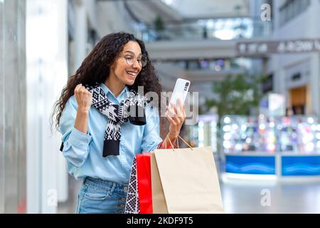 Porträt einer glücklichen Shopper-Frau, hispanische Frau im Einkaufszentrum, nutzt Smartphone, durchsucht Online-Rabatte und -Verkäufe, hält Hand hoch, feiert empfangene Angebote, gewinnt. Stockfoto