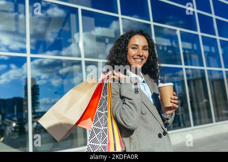Porträt einer glücklichen Shopper-Frau, einer hispanischen Frau, die vor dem großen Supermarkt die Kamera anschaut und lächelt, mit bunten Taschen und einer Tasse heißem Getränk. Stockfoto