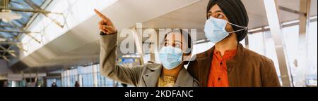 Indisches Paar mit Gesichtsmasken, das sich umarmt und mit dem Finger in den Innenraum des Flughafens zeigt Stockfoto