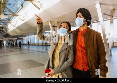 Indisches Paar mit Gesichtsmasken, das sich umarmt und mit dem Finger in den Innenraum des Flughafens zeigt Stockfoto