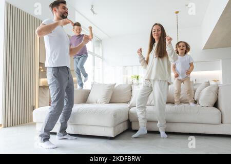 Lustige aktive Familie aus vier jungen erwachsenen Eltern und niedlichen kleinen Kindern, die im Wohnzimmer zusammen tanzen, sorglose kleine Kinder mit Mama Papa ha Stockfoto