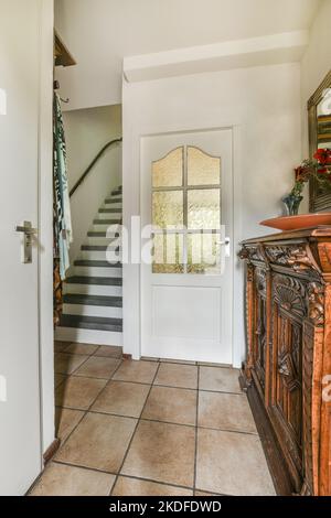 Vintage-Schrank mit Dekorationen und Spiegel in der Nähe offene Wohnzimmertür und Treppe im Flur des Hauses Stockfoto