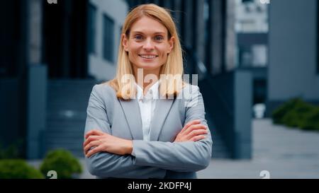 Erfolgreiche glückliche junge Geschäftsfrau geht auf der Straße selbstbewusst stolze Frau professionelle Büromanagerin posiert mit gekreuzten Armen im Freien zu suchen Stockfoto