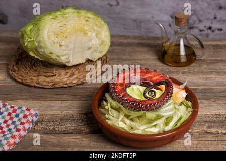 Tonschale mit in Streifen geschnittenen Kohlblättern und einem Stück getrockneten Tintenfisch, der auf dem Grill auf einem rustikalen Holztisch mit einer Öldose und einem pi gekocht wird Stockfoto