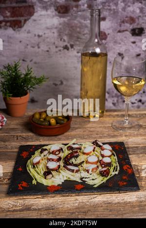 Gegrillter, getrockneter Oktopus auf einem Bett aus zerkleinerten Kohlblättern auf einem Steinteller, neben einigen Oliven, einer Flasche Wein und einem Glas Weißwein. Stockfoto