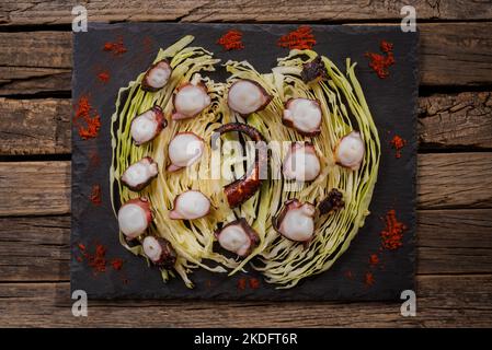 Getrocknete Tintenfischkeule auf dem Grill gekocht und in Stücke geschnitten, auf einem Bett aus zerkleinerten Kohlblättern auf einem Steinteller, auf einem rustikalen Holztisch. Stockfoto