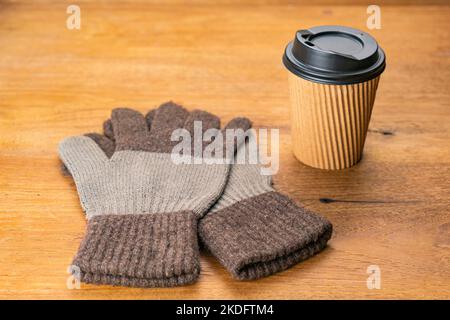 In der Wintersaison ein Paar Stoffhandschuhe und eine schwarze Kunststoffkappe aus Papier zum Mitnehmen einer Tasse heißen Kaffees auf einem Holztisch. Stockfoto