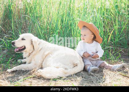 Ein wunderschönes Baby im Anzug mit Hosenträgern sitzt mit dem Hund im Gras Stockfoto