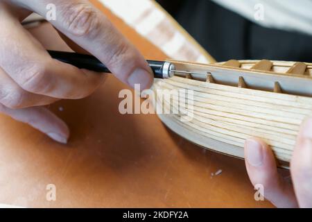 Hände des Mannes, der überschüssigen Klebstoff von Sperrholzdetails für Schiffsmodell mit Gebrauchsmesser entfernt. Prozess des Bauens Spielzeug Schiff, Hobby, Handwerk. Tabelle mit Stockfoto